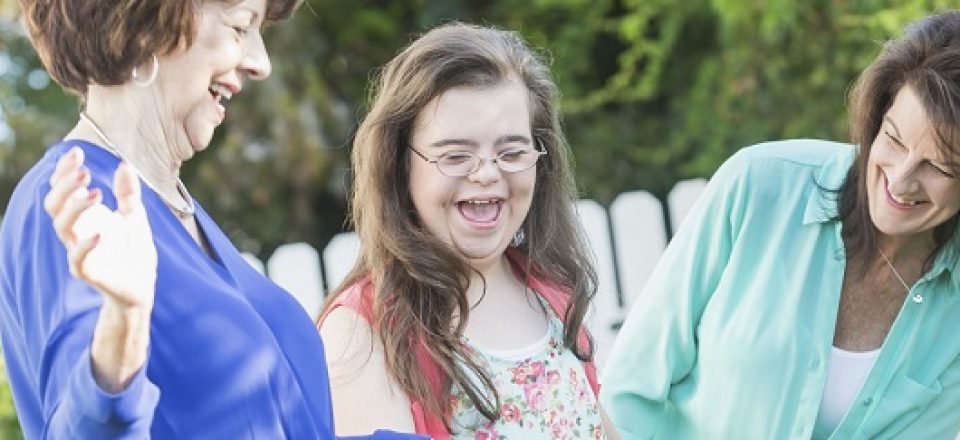 A teenager girl with down syndrome, laughing and having fun with her mother and grandmother outdoors in the back yard.  She is standing in the middle holding her grandmother's hand.
