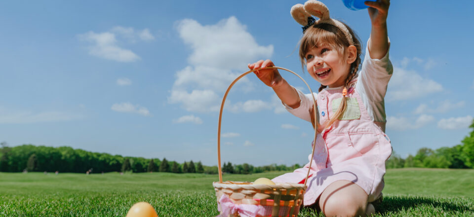 Little girl wearing rabbit ears hoop picking up and putting colorful toy eggs in a basket outdoor. Easter tradition for children.